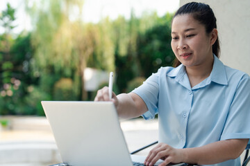 Asian woman using digital stylus on laptop. Woman remote working, creative design, online education and communication in comfortable. Freelance woman lifestyle with nature inspired creativity.