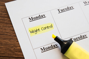 Weight control reminder highlighted on weekly planner with yellow marker lying nearby on wooden desk. handwritten note emphasizes health, fitness, self-discipline in maintaining balanced lifestyle