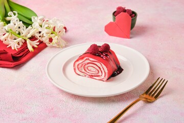 Sliced ​​sweet roll of thin red pancakes with cream, decorated with chocolate and raspberries, on a white plate on a pink background. Valentine's Day.