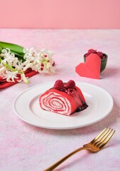 Sliced ​​sweet roll of thin red pancakes with cream, decorated with chocolate and raspberries, on a white plate on a pink background. Valentine's Day.