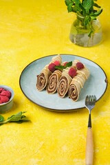 Thin crepes with poppy seed filling, decorated with raspberries and white chocolate, on a gray plate on a yellow concrete background.