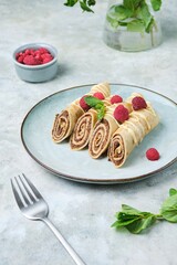 Thin crepes with poppy seed filling, decorated with raspberries and white chocolate, on a gray plate on a yellow concrete background.