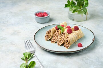 Thin crepes with poppy seed filling, decorated with raspberries and white chocolate, on a gray plate on a yellow concrete background.