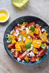 Salad with red tomatoes, grilled corn and cheese in a blue bowl, vertical shot on a brown granite background, high angle view