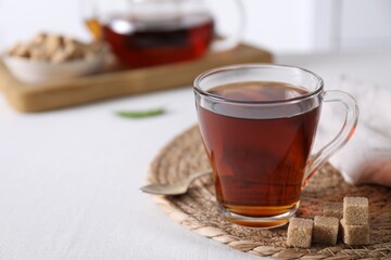 Aromatic black tea in cup and brown sugar cubes on white table, closeup. Space for text