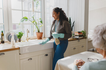 Rare view of elderly lonely woman talking to young african american female helping volunteer while she cleaning her kitchen sink in blue raisin gloves, spraying and mopping dirt from surfaces