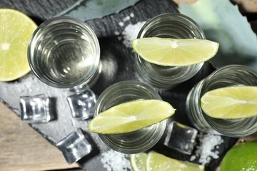 Tequila shots with lime slices, salt, ice cubes and agave leaf on wooden table, flat lay
