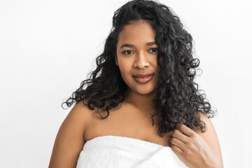 Spa, resort and sauna concept. Adorable black plus size woman standing against white copy space background in bath towel, feeling fresh and relaxed after taking shower with organic cosmetic products
