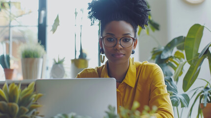 Black woman working on laptop surrounded by plants in office.