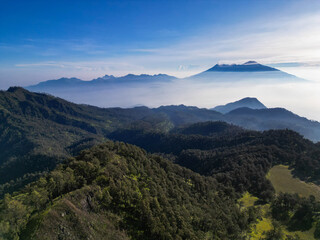 Aerial view drone natural beauty of green hills and grasslands adorns the foreground. Mount Arjuno Welirang towers over a thin blanket of mist. The sunlight casts a long shadow effect on the hillside.