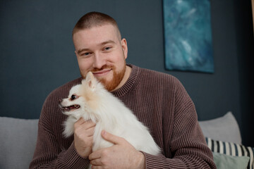 Portrait of smiling man holding a fluffy white Pomeranian while sitting on a couch. Man is in a...
