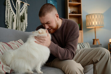 Man cuddling white Pomeranian dog on sofa in cozy living room filled with soft light. Decorative elements, like woven wall hanging and lamp, adding warmth