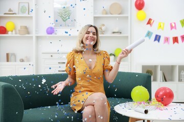 Happy woman blowing up confetti popper on sofa in room decorated for party
