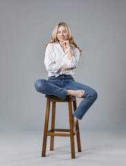 Smiling woman in stylish jeans sitting on stool against grey background