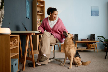 Woman sitting at desk in cozy home office, smiling at attentive dog with bookshelf nearby. Room decorated with plants and candle, creating inviting atmosphere