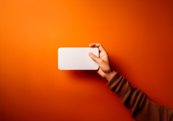 Obraz premium A hand with a brown sleeve holds a white rectangular object against an orange backdrop. The blank object is held horizontally in a simple studio shot with a focus on the hand and the object.