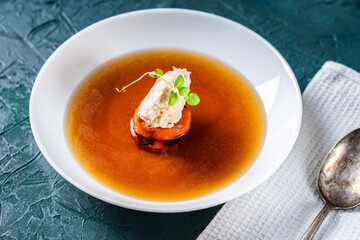 Soup or consomme, thick meat broth with carrots, chicken dumplings and microgreens, served in a white plate on a dark green background with a napkin and a spoon.