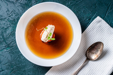 Soup or consomme, thick meat broth with carrots, chicken dumplings and microgreens, served in a white plate on a dark green background with a napkin and a spoon. View from above