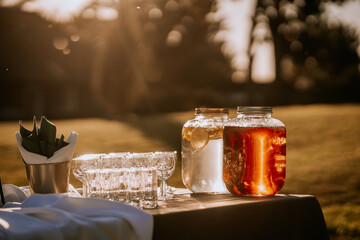 Outdoor table with jars of water and iced tea, glassware, and napkins, bathed in warm golden sunlight during a serene sunset in nature.