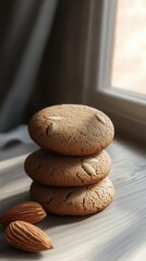 A stack of almond cookies sits on a wooden surface near a window