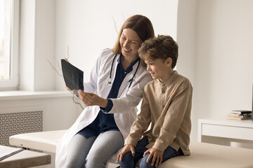 Obraz premium Useful radiation. Caring attentive young female nurse doctor pediatrician surgeon radiologist make x ray diagnostics study radiograph together with interested young patient boy describe picture to kid