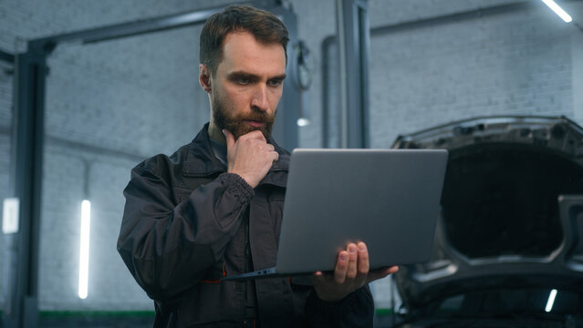 Serious focused Caucasian man guy technician auto mechanic engineer check repairing car problem computer diagnostics electric automobile engine with laptop checking maintenance in garage vehicle shop