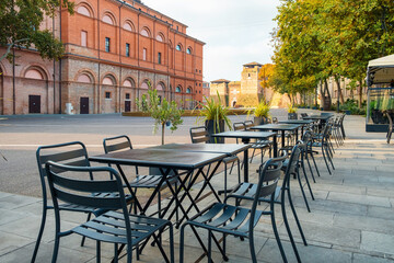 Outdoor cafe with tables and chairs on paved terrace in old town square in Rimini, Italy, with view of Castel Sismondo and historic Renaissance fortress. Italian piazza with sidewalk restaurant