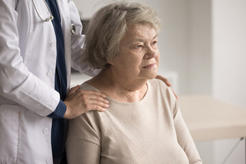 Fototapeta premium Older female retiree patient of nursing home for elderly people sit on chair look away feel support of caregiver nurse hugging her shoulders from back. Medical aid for aged persons concept. Copy space