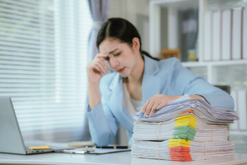 Tired Asian businesswoman sitting at desk working, pile of documents and looming deadline. Stress and fatigue visible on her face as she works on laptop.