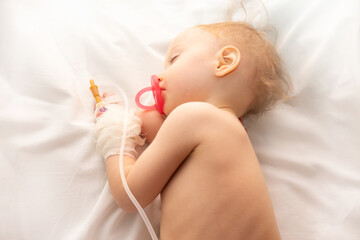 Babygirl sick and sleeping on patient bed at the hospital. Saline intravenous or (IV)drip in baby patient hand. Poor baby looks tired and exhausted.IV fluid replacement or correcting the dehydration