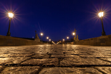 A cobblestone bridge stretching into the distance at night, illuminated by glowing street lamps and...
