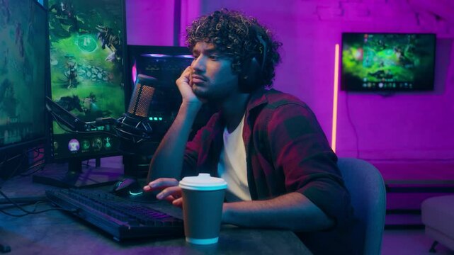 Sleepy bored Arabian Indian Latino man playing video game cyber sport streamer gamer lazy guy drinking coffee tired male e-sport online computer home addict studio exhausted sleeping boredom streaming