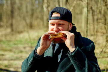 Man enjoying a snack outdoors in a forest during a sunny day while wearing a black cap and coat