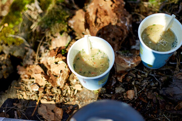 A group of friends relaxes outdoors, sipping warm drinks from paper cups. Bright sunlight illuminates their gathering beside a kettle on mossy ground.