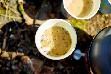 A group of friends relaxes outdoors, sipping warm drinks from paper cups. Bright sunlight illuminates their gathering beside a kettle on mossy ground.