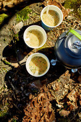 A group of friends relaxes outdoors, sipping warm drinks from paper cups. Bright sunlight illuminates their gathering beside a kettle on mossy ground.