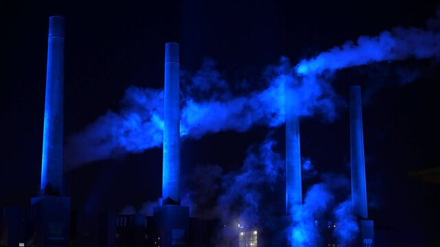 Smoking factory chimneys at night, illuminated by flashing blue spotlights. Wolfsburg, Germany