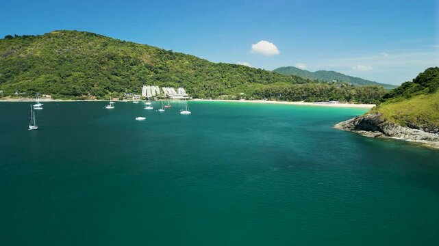 Drone view of Nai Harn Beach with white yachts anchored in turquoise waters, surrounded by lush hills and a tropical coastline in Phuket, Thailand.