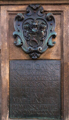 A close-up shot of a weathered stone plaque featuring an ornate crest above an inscription in raised text, mounted on a textured stone wall.