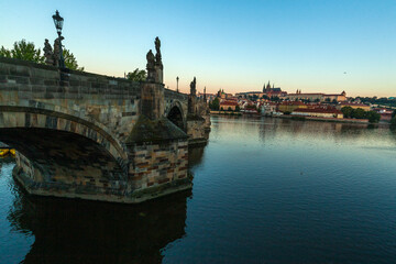 A stone bridge with statues lining its edge spans a wide river, offering a distant view of a cityscape with red-roofed buildings and a large complex under a clear sky at twilight.