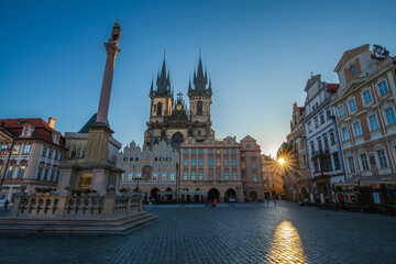 Fototapeta premium A cobblestone square featuring a tall monument with a statue, a grand gothic church with spires, and ornate buildings lining the square under a clear sky with sunlight.
