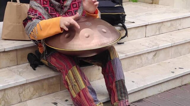A young guy with dreadlocks and a beard plays a percussion musical instrument Hang in the city on the street.