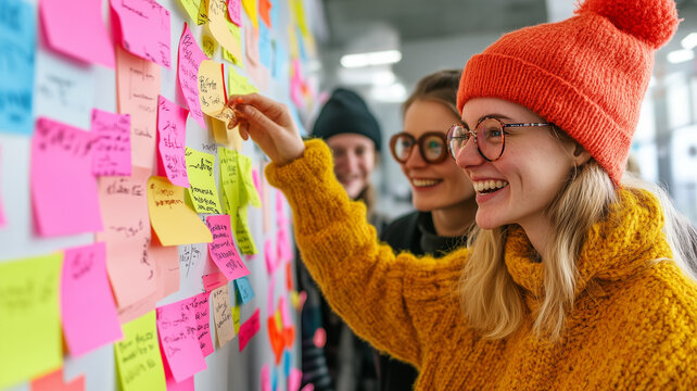 Creative brainstorming session with colorful sticky notes on wall. Three young women collaborate, sharing ideas and smiling, showcasing teamwork and innovation