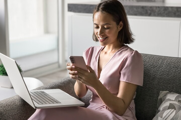 Young pretty woman sits comfortably on sofa with laptop, smile, holding smartphone, enjoy remote...