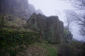 An old, moss-covered stone house with castle-like features, nestled against a rocky hillside in a misty, eerie landscape. The foggy atmosphere and overgrown surroundings create a mysterious and abando