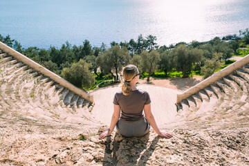 woman sits on ancient amphitheater ruins, gazing at the vast blue sea. This serene moment captures...