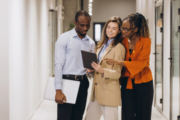 Professional diverse colleagues reviewing project details together, walking workplace hallway with shared digital tablet