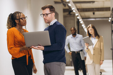 Multi ethnic business team working together using laptop and digital tablet, discussing projects in office corridor