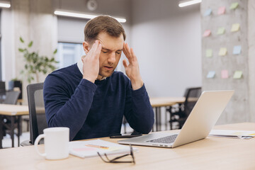 Businessman suffering workplace stress, holding head near laptop in contemporary workspace, embodying professional burnout