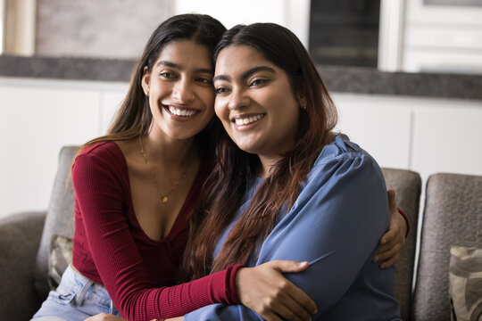 Two attractive Indian women sitting close together on sofa in cozy modern living room, cuddling, feeling sibling love, showing mutual support, enjoy shared moments. Closeness and trust between sisters